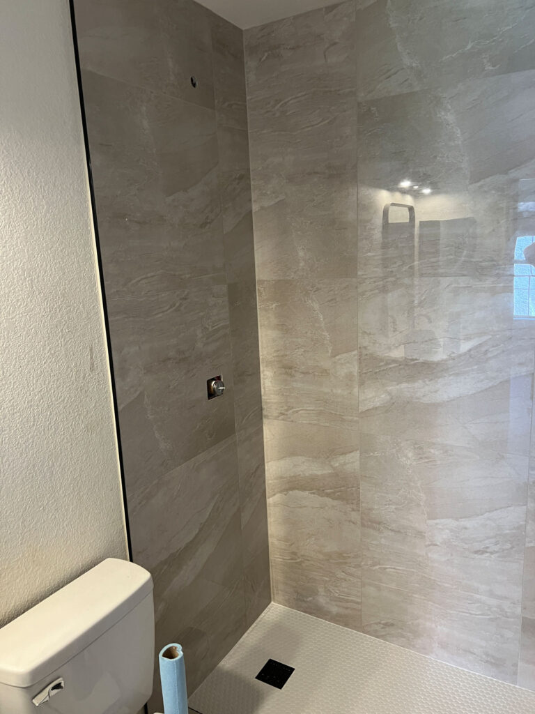 A modern bathroom with light beige tiled walls, a white toilet, a shower area with a drain, and a roll of blue paper towel on the toilet tank creates a fresh bath atmosphere.