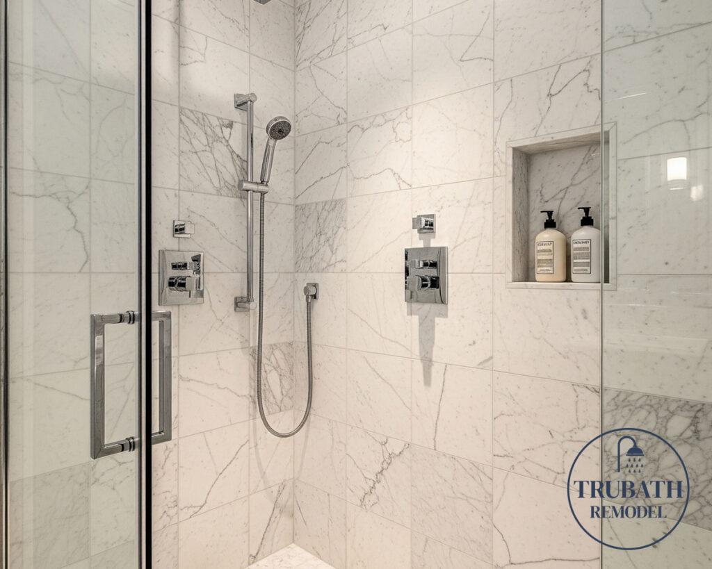 Modern shower with white marble tile, glass door, built-in niche holding two bottles, and chrome fixtures. Trubath Remodel logo is visible in the bottom right corner.