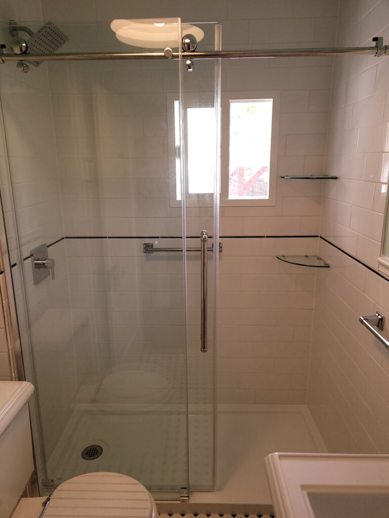 Modern bathroom remodel in Phoenix featuring a glass-enclosed shower, white subway tile walls, a small window, two glass corner shelves, and sleek metal fixtures. Part of a toilet and sink are visible.
