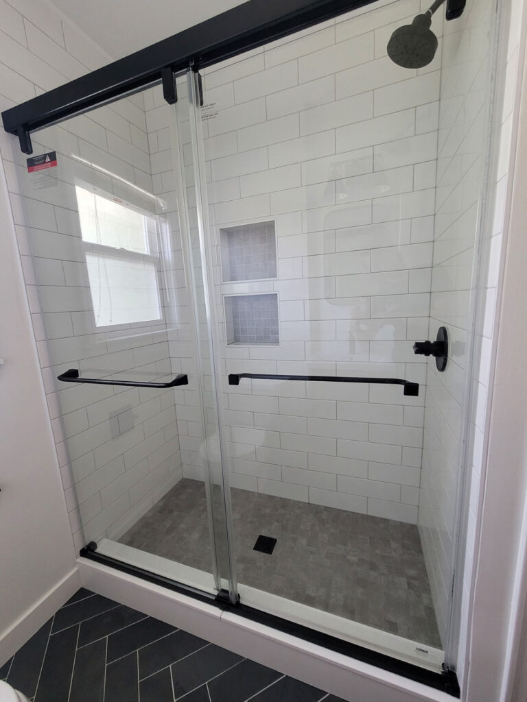 Modern shower with sliding glass doors, white subway tile walls, dark fixtures, built-in shelves, and a gray tiled floor—perfect for your next bathroom remodel in Phoenix.