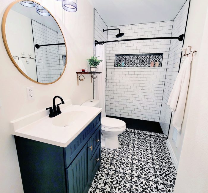 Modern bathroom remodel with a navy vanity, round mirror, black fixtures, bath and shower, built-in shelf, and black-and-white patterned floor tiles.