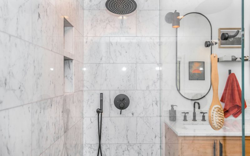 Modern bath with marble shower walls, black fixtures, a rain showerhead, vanity with rectangular mirror, and a hanging wooden bath brush. A red towel hangs on the wall, creating a stylish wet space by Trubath.
