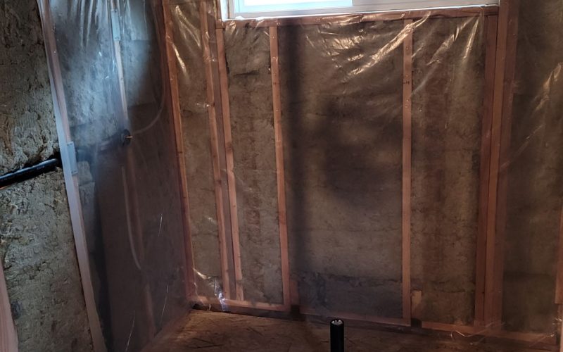 Room under construction for a Trubath remodel, featuring exposed wall insulation, plastic sheeting, a small horizontal window, plywood floor, and a black pipe protruding from the floor.
